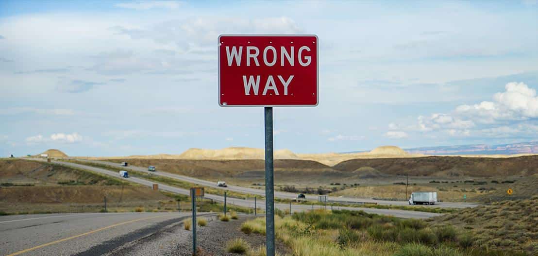 A red "Wrong Way" sign stands beside a highway in a vast, open landscape with rolling hills under a partly cloudy sky. A few vehicles and long roads stretch into the distance.