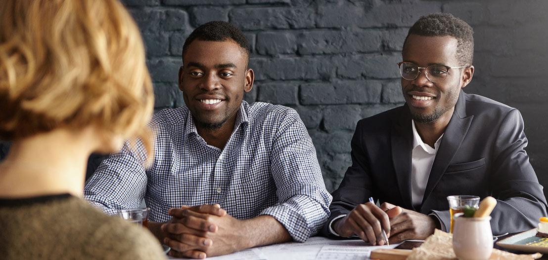 Two men sit across from a woman at a table, engaged in conversation. One wears a checkered shirt, the other a suit jacket and glasses. They appear to be in a casual meeting with drinks and papers, conducting a structured interview in the softly lit room.