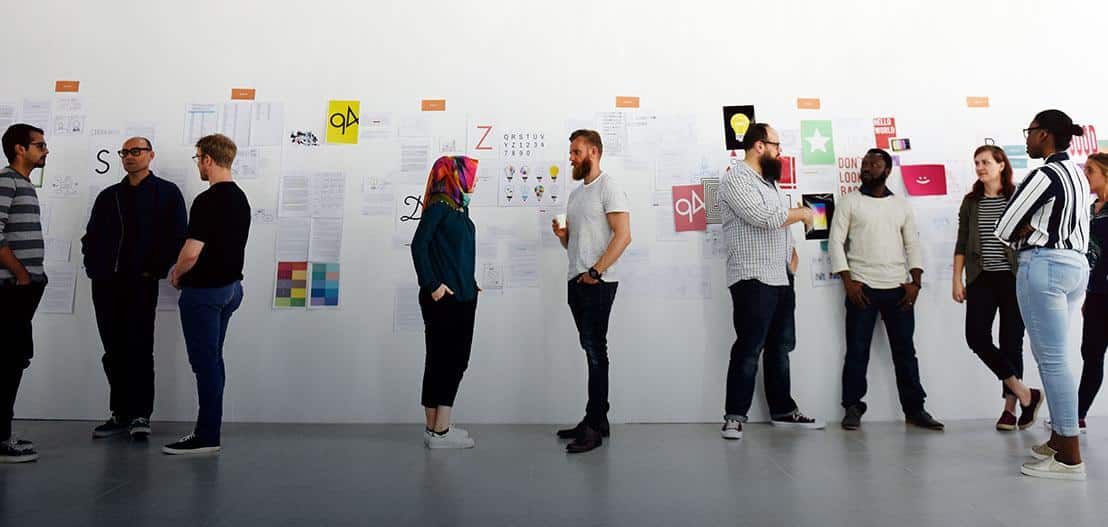Nine people stand and chat in front of a large white wall covered with various colorful posters and papers. They appear to be in a casual discussion, possibly a creative meeting or workshop, in a bright, minimalistic room.