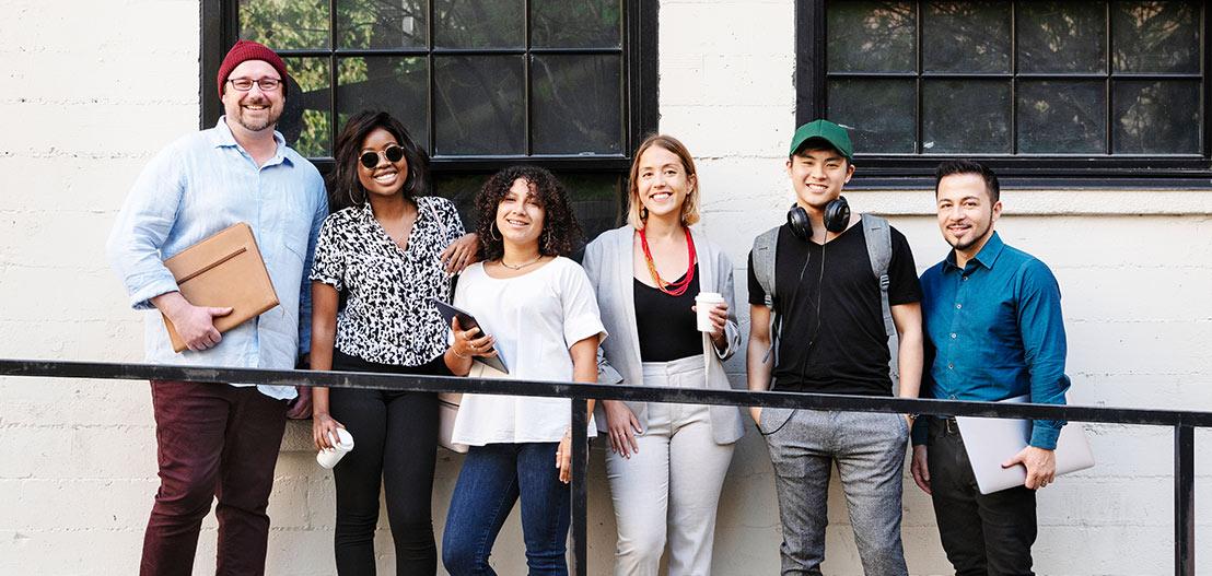 A diverse group of six people standing in front of a building with windows. They are smiling and casually dressed, holding various items like a laptop, coffee cup, and headphones. It appears to be a sunny day.