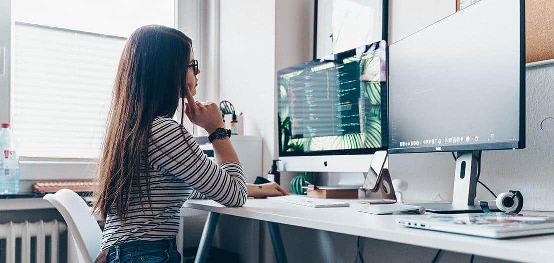 A woman with long hair, wearing glasses and a striped shirt, sits at a desk working on a computer with dual monitors displaying code. The room is bright with natural light from a nearby window. A water bottle and various desk items are visible.