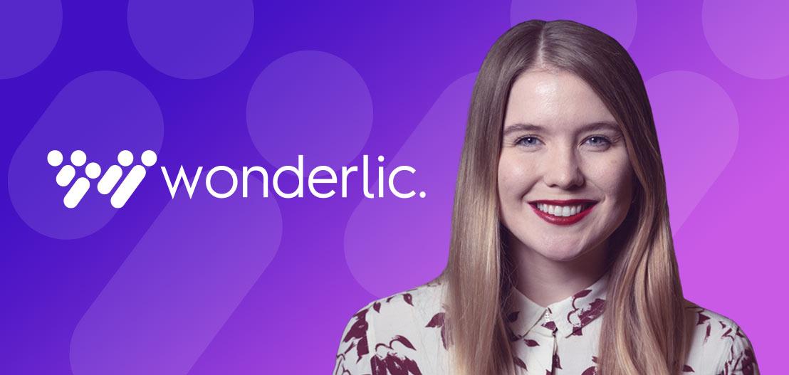 A smiling woman with long, light brown hair stands in front of a purple background. She is wearing a patterned shirt. The word "wonderlic." appears on the left side of the image in white text.
