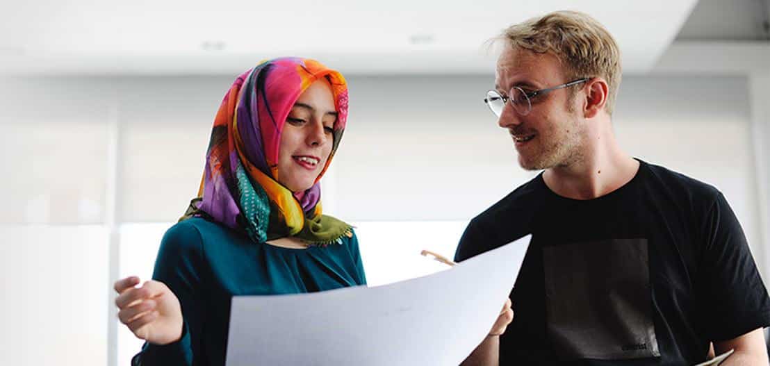 A woman wearing a colorful hijab holds a document and gestures while talking to a man in glasses and a black shirt. They appear to be discussing the contents of the document in a bright room.