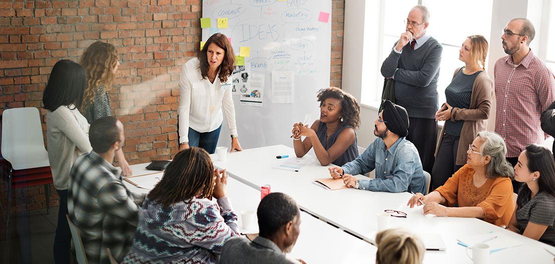 A diverse group of people engaged in a collaborative meeting around a long table, focusing on soft skills. A woman stands, addressing the group, which includes men and women of various ages and ethnicities, while notes and ideas are visible on the board behind her.