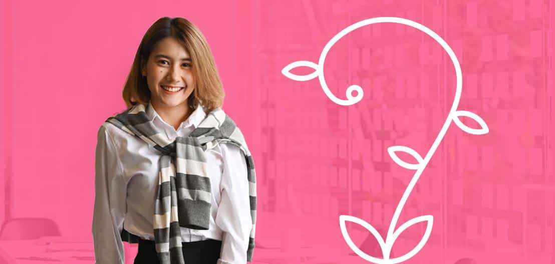 A woman with shoulder-length hair smiles at the camera. She wears a white shirt and a striped sweater draped over her shoulders. The background is pink with a white vine design.
