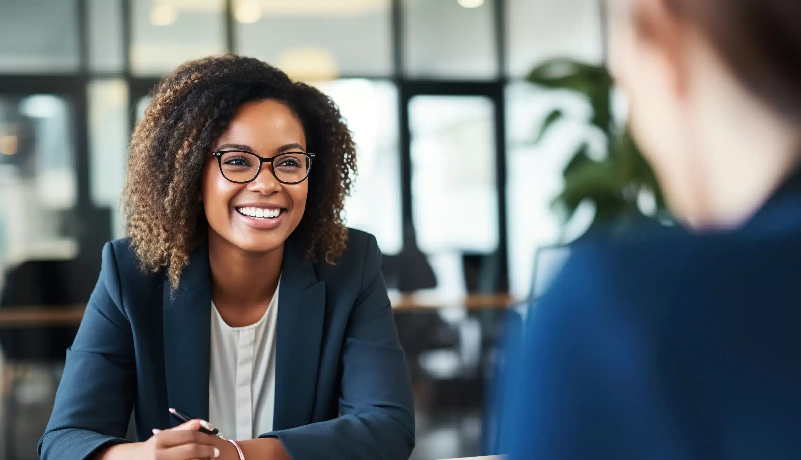 A woman with curly hair and glasses, wearing a dark blazer, smiles while sitting at a desk discussing team effectiveness with another person in a modern office setting.