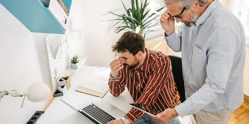 Two men at a desk look stressed; one is sitting with his hand on his face, while the other stands holding a tablet. A laptop, notebook, and plant are on the white desk in a home office setting.