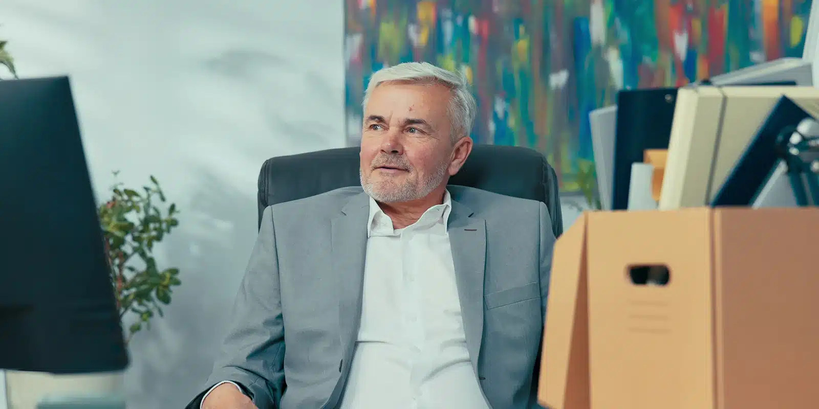 An older man with gray hair and a beard, wearing a gray suit, sits in an office chair near a cardboard box and computer, appearing thoughtful, perhaps contemplating voluntary vs involuntary turnover. Colorful abstract artwork hangs on the wall behind him.