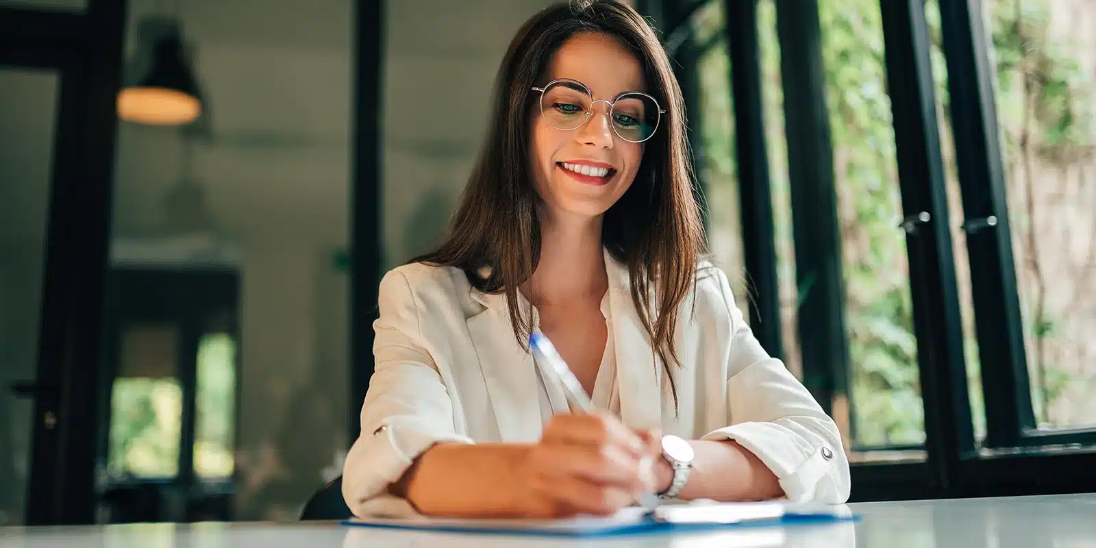 A woman with long brown hair and glasses smiles while writing on paper at a desk, wearing a white blazer. She appears to be completing an employee engagement survey as she sits in a bright room with large windows and greenery outside.