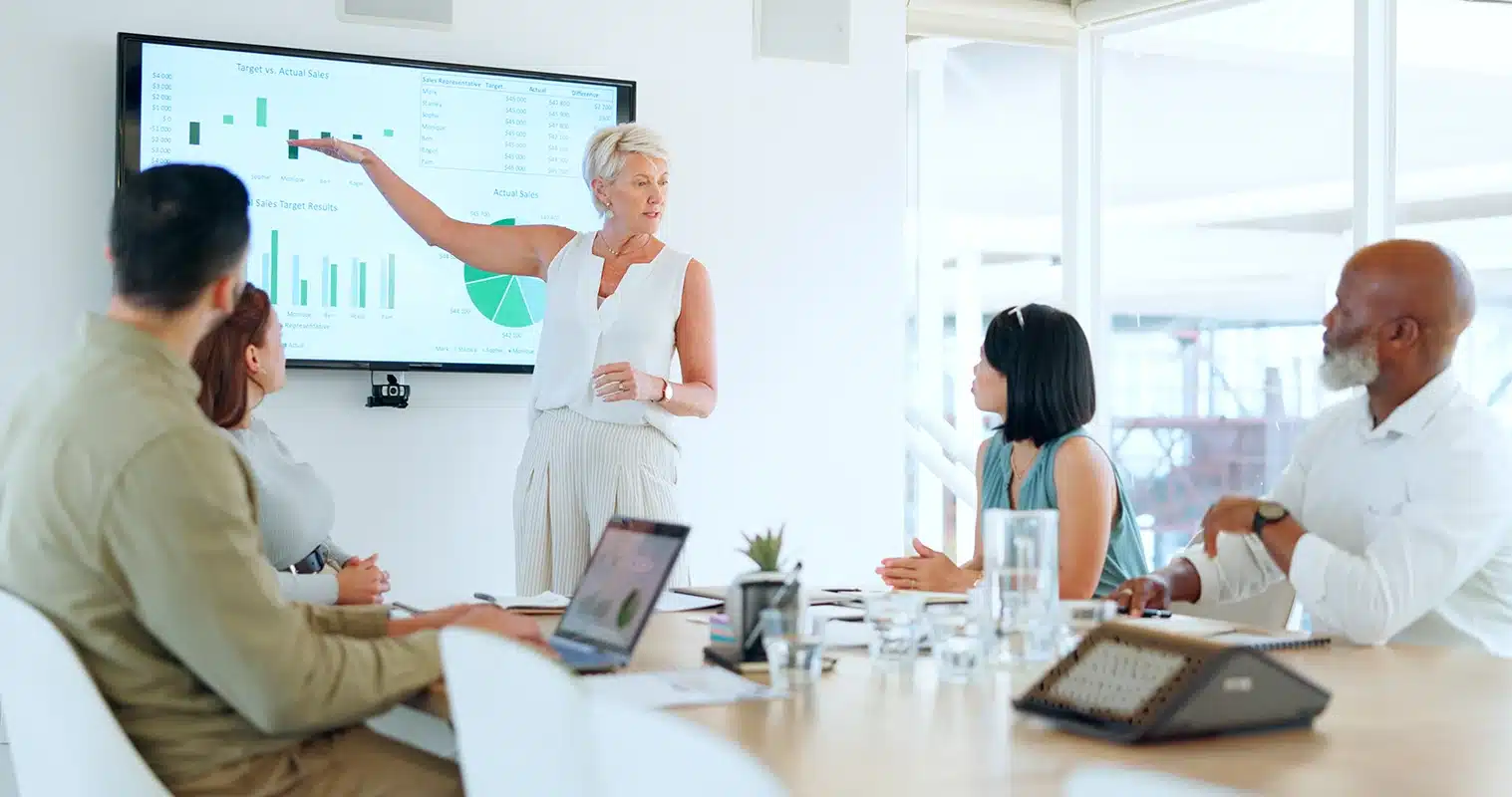 A woman stands and points to a presentation slide with charts on a screen, highlighting employee turnover rate benchmarks, while four colleagues sit around a conference table listening attentively.