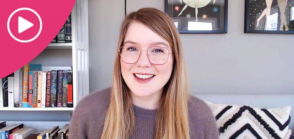 A person with long hair and glasses smiles at the camera. They are indoors, with shelves of books to one side and framed pictures on the wall. A pink play button icon is in the top left corner.