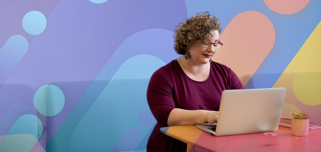 A woman with curly hair and glasses is smiling while using a laptop. She is seated at a colorful table with a small plant on it. The background features a vibrant, abstract mural with overlapping shapes in purple, orange, and blue.