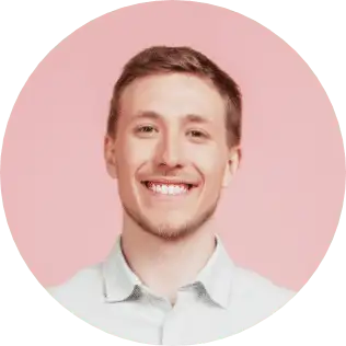 A man with short light brown hair is smiling broadly against a pastel pink background. He is wearing a light-colored button-up shirt and inviting you to get started with a Wonderlic demo. The image is circular.