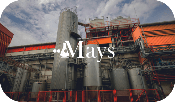 Industrial building with large silos and machinery under a cloudy sky. Red and orange accents mark parts of the structure. Branded text "Mays" with a magnifying glass and beaker icon overlay, highlighting the site's rigorous hiring assessment process.