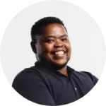 A person with short hair smiles warmly at the camera, wearing a dark shirt. The background is plain white.