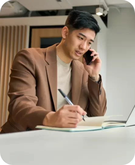 A person in a brown blazer is sitting at a table, talking on a cellphone while writing in a notebook. A laptop is open in front of them. The background features modern office decor with wood paneling.