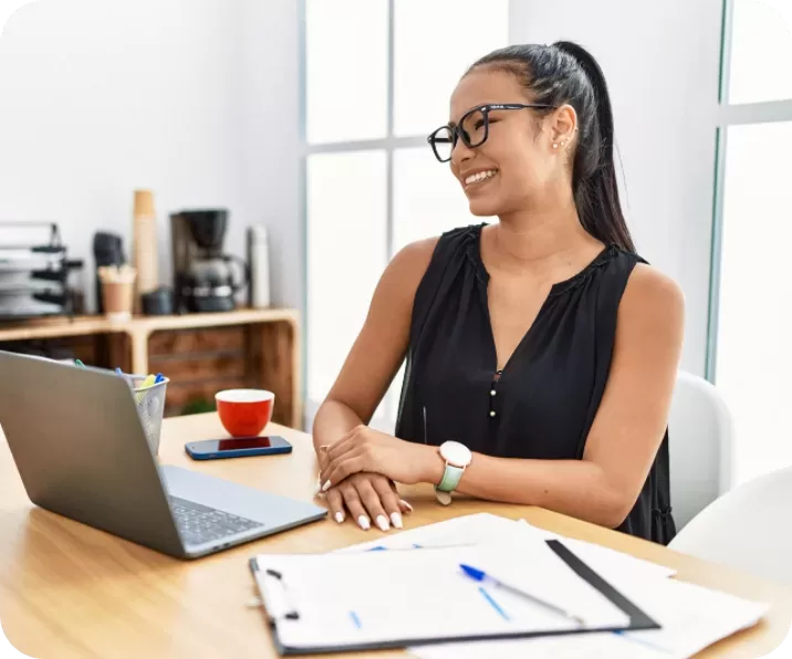 A woman wearing glasses and a black sleeveless top sits at a desk with a laptop, smiling. On the desk are a red cup, a phone, and some documents. The background features office supplies and a coffee maker.