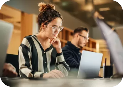 A woman with glasses and curly hair tied up is focused on her laptop at a desk in a modern office. She is wearing a striped blouse. In the background, a man is also working on a laptop. The office is well-lit and has a contemporary design.