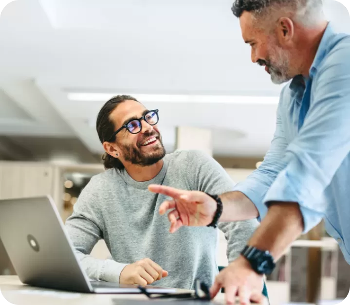 Two men in a bright office setting are interacting. One is seated, smiling, and wearing glasses while looking at the other, who is standing, pointing, and smiling back. A laptop is open in front of them on a table.