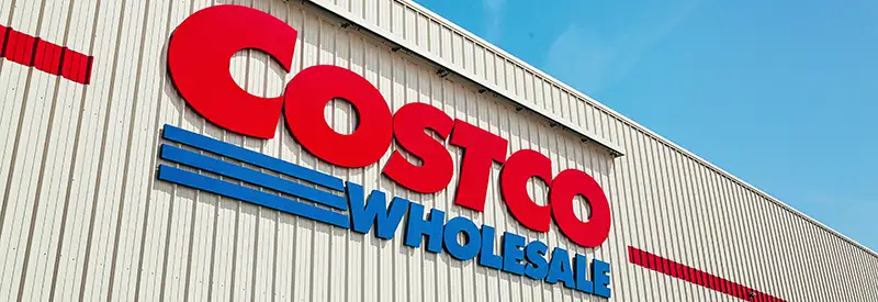 Large red and blue "Costco Wholesale" sign on the exterior wall of a beige warehouse building, viewed from below with a clear blue sky—a testament to the team dynamics that support such a thriving retail giant.