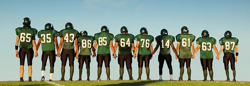 Eleven American football players in green jerseys stand side by side outdoors, holding hands on a grassy field under a clear blue sky, symbolizing strong team dynamics as they face the game together.