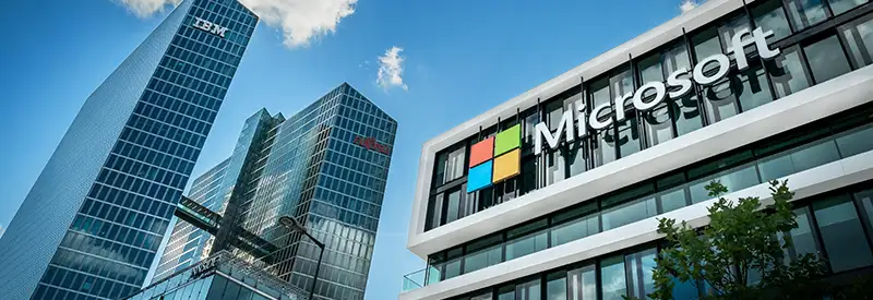 Modern office buildings under a blue sky showcase prominent IBM and Microsoft logos, symbolizing innovative team dynamics. The Microsoft logo stands out in the foreground, with IBM displayed on a taller building in the background.