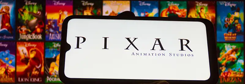 A smartphone displays the Pixar Animation Studios logo, held in front of a screen showing various colorful Disney animated movie posters, highlighting the team dynamics behind these beloved films.