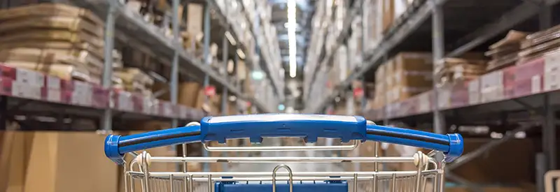 A shopping cart is pushed down a wide aisle in a large warehouse store, where team dynamics are at play as staff organize tall shelves stacked with various boxes and goods on both sides.