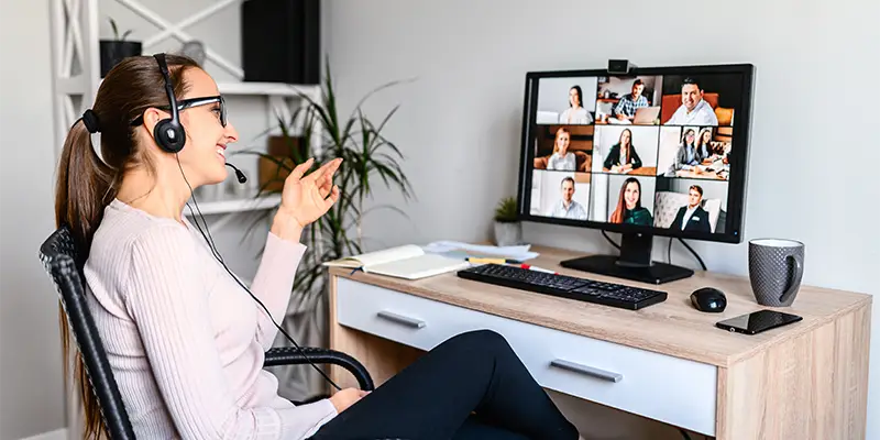 A woman wearing a headset sits at a desk, smiling and participating in a video conference with multiple people displayed on her screen, reflecting positive team dynamics in the workplace. A notebook, coffee mug, and phone sit on the desk.