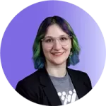 Person with short, wavy hair dyed blue and green, wearing glasses, a gray shirt with a white design, and a black blazer, smiling in front of a purple gradient background—a look that reflects confidence and effective coaching expertise.