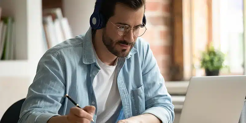 A man wearing headphones and glasses sits at a desk, writing with a pen while looking at a laptop. He appears focused, maintaining assessment integrity, with shelves and a window in the background.