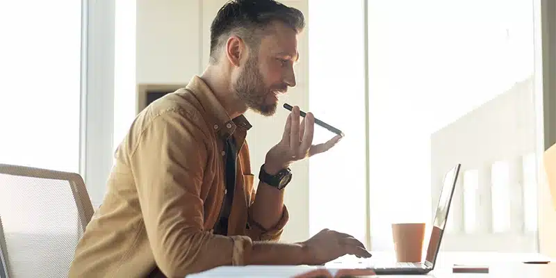 A man sits at a desk using a laptop, speaking into his smartphone. In the bright office space with sunlight streaming through large windows, he appears to be utilizing AI at work. A cup sits on the desk beside him.