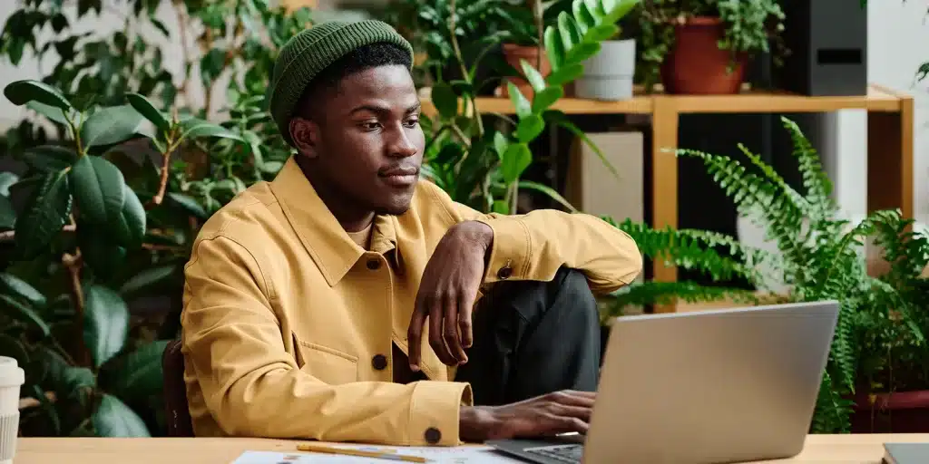 A person in a yellow jacket and green beanie sits at a desk with a laptop, surrounded by green plants, appearing focused on their work—embodying the Gen Z AI-driven approach to productivity.