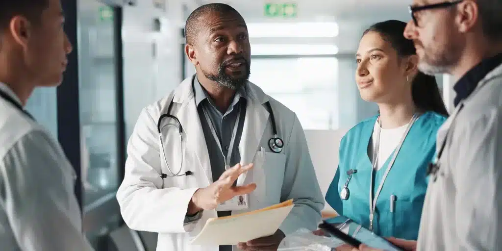 A doctor in a white coat conducts employee onboarding with a group of medical colleagues in a hospital hallway.