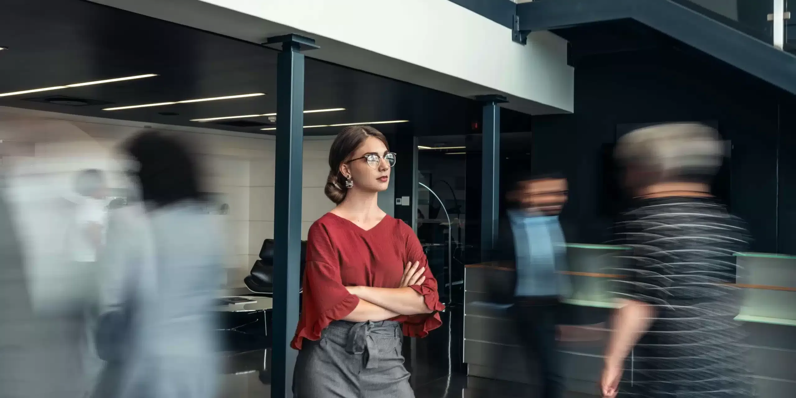 A woman in a red blouse and gray pants stands confidently with arms crossed in a modern office building, highlighting her strong role amid the fast-paced team dynamics as people around her blur by, suggesting movement and busyness.