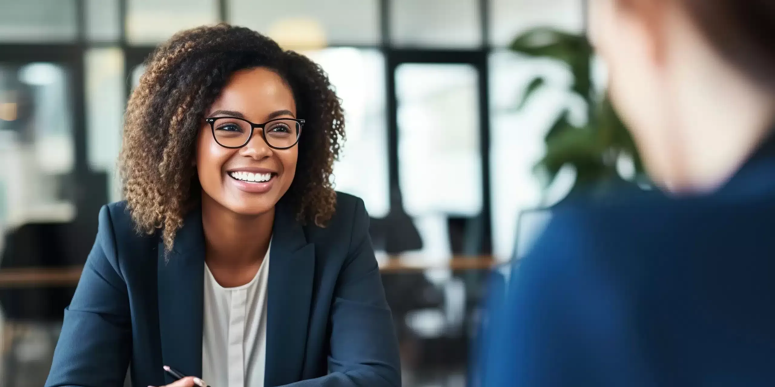 A woman with curly hair and glasses, wearing a dark blazer, smiles while sitting at a desk discussing team effectiveness with another person in a modern office setting.
