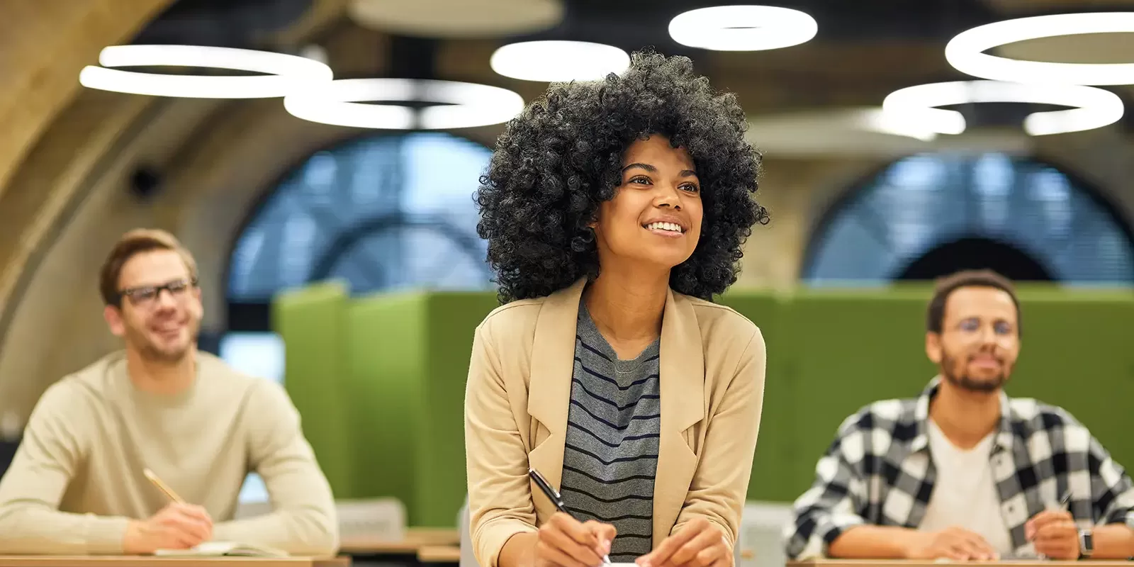 Three people sit at desks in a modern classroom. A woman in the center, smiling, writes in a notebook while preparing for Wonderlic certification. Two men on either side are also engaged, with blurred green partitions and circular lights behind them.