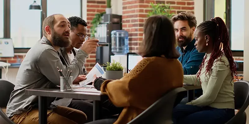 Five people sit around a table in a modern office, engaged in discussion. Papers, laptops, and a small plant are on the table as group dynamics unfold. Large windows and brick walls are visible in the background.