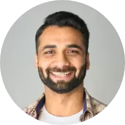 A person with short dark hair and a beard is smiling at the camera during an employee assessment. They are wearing a plaid shirt over a white T-shirt, set against a plain, light-colored background.